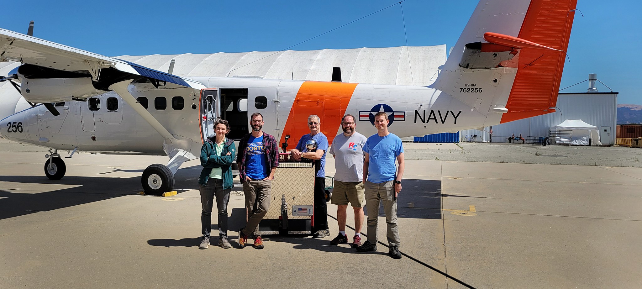 Five people stand centered in the image in front of an aircraft. They are smiling at the camera and wearing casual outdoor type clothes. The aircraft is facing left in the image