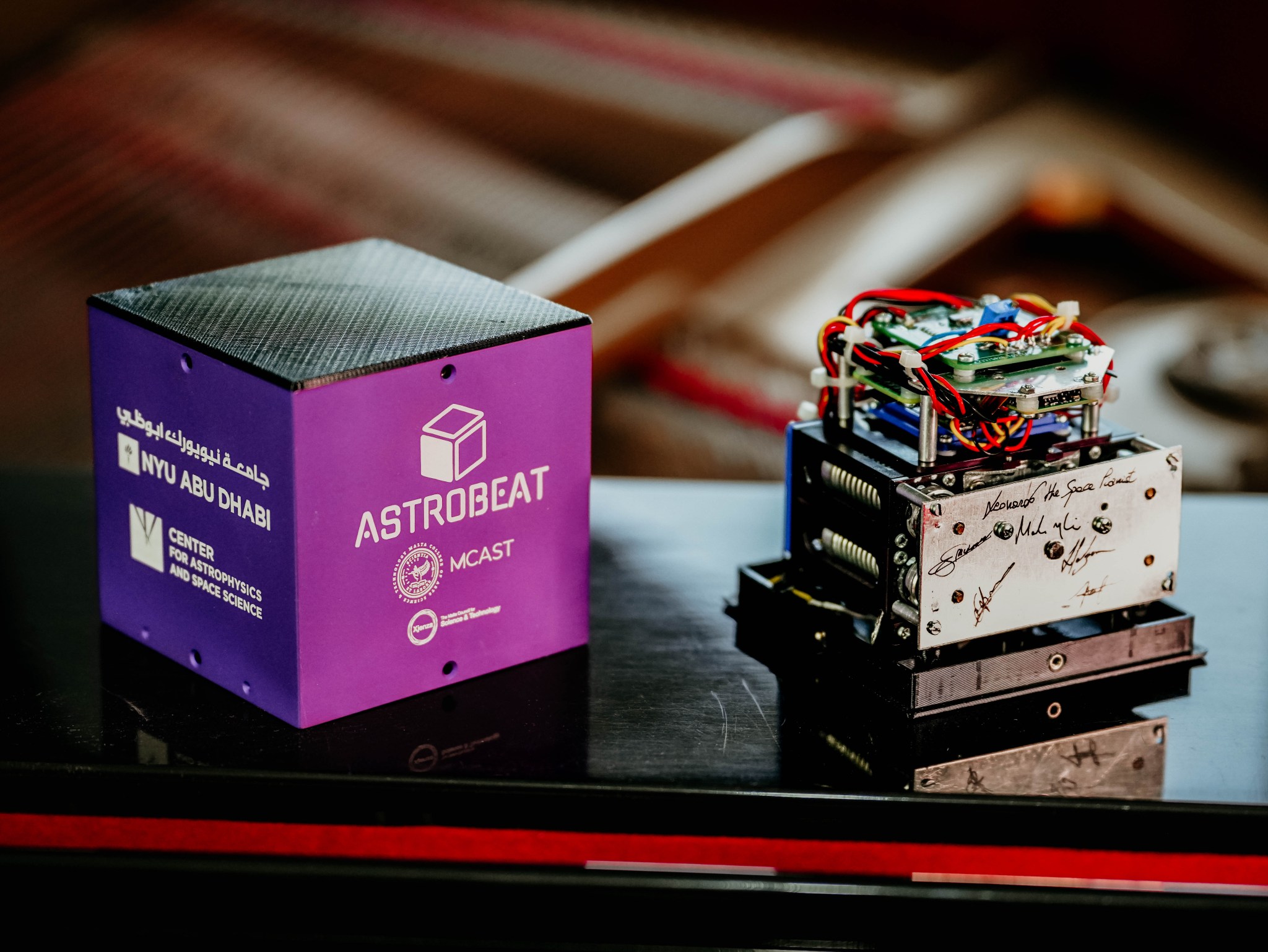 Two items sit on a shiny black surface. On the left is a small cube with a silver top and purple sides labeled “NYU Abu Dhabi” on one side and “MCAST” on the other. On the right is the device that fits inside the cube. It has a snarl of colored wires around a metal plate above a box that holds the samples with two springs visible on its side.