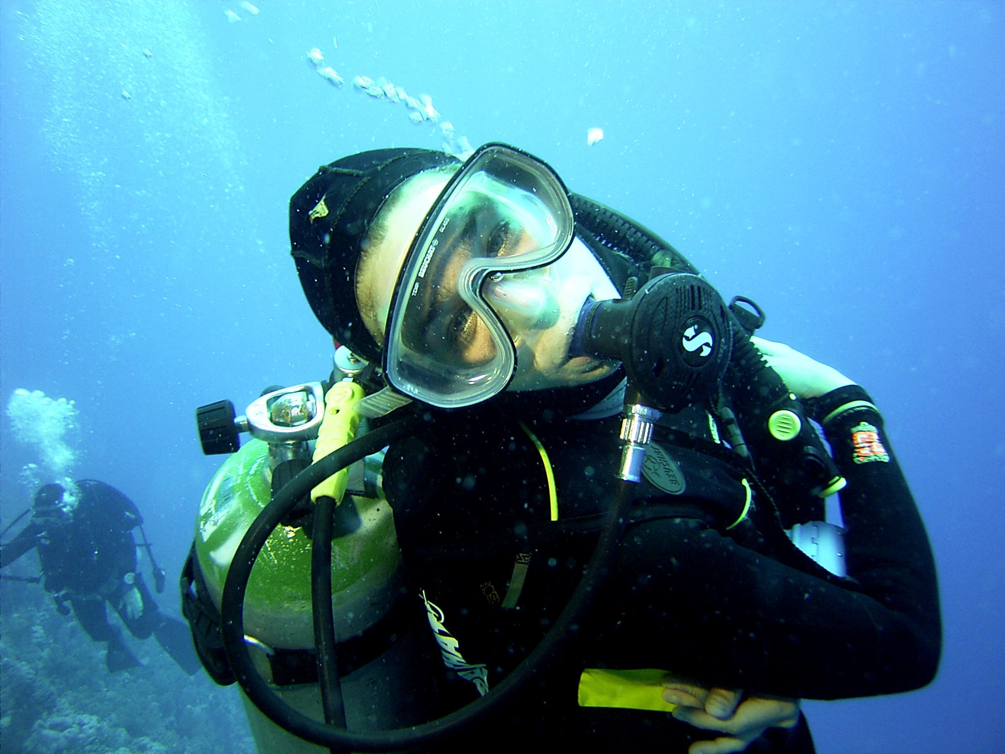 A photo of  Meloë Kacenelenbogen looking at the camera, underwater, wearing full scuba diving gear including a mask, oxygen tank, and snorkel. Another diver is visible in the background. 