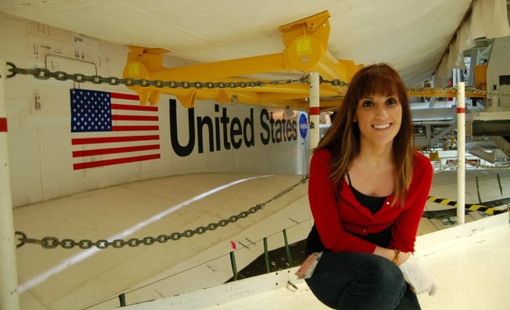 A woman sits in front of a large NASA spacecraft component. She is wearing a red cardigan over a black top and jeans.