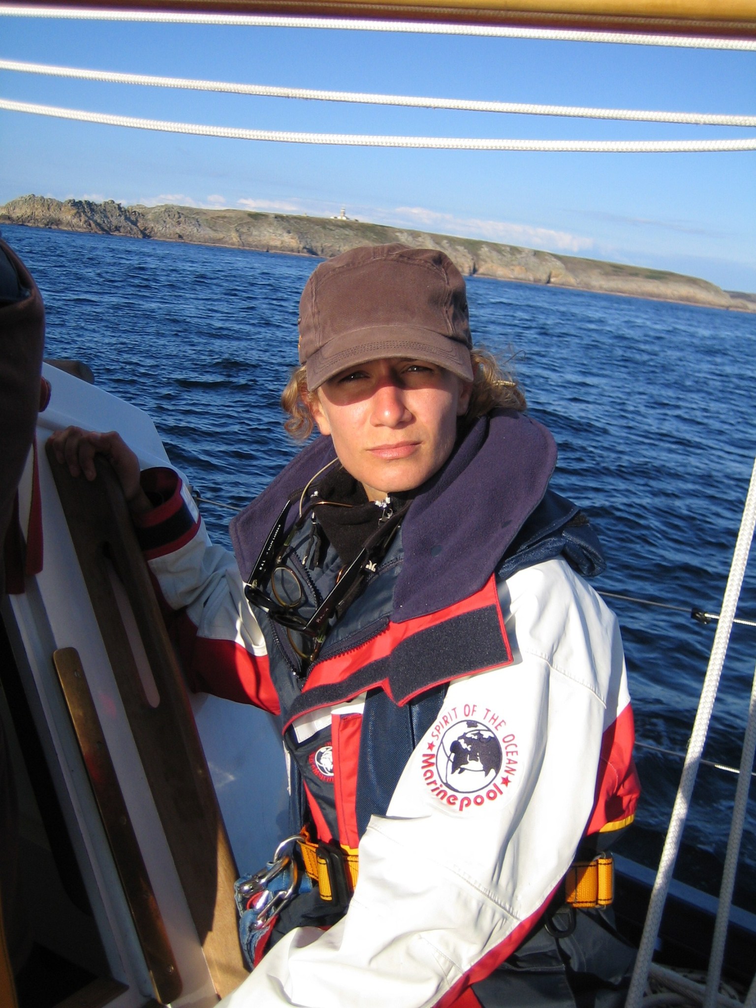 Meloë S. Kacenelenbogen looks at the camera on a boat with blue water and land in the background. She is wearing a black hat and a red, white, and blue jacket that has a logo with a globe and the words "Spirit of the Ocean, Marinepool" on the sleeve. 