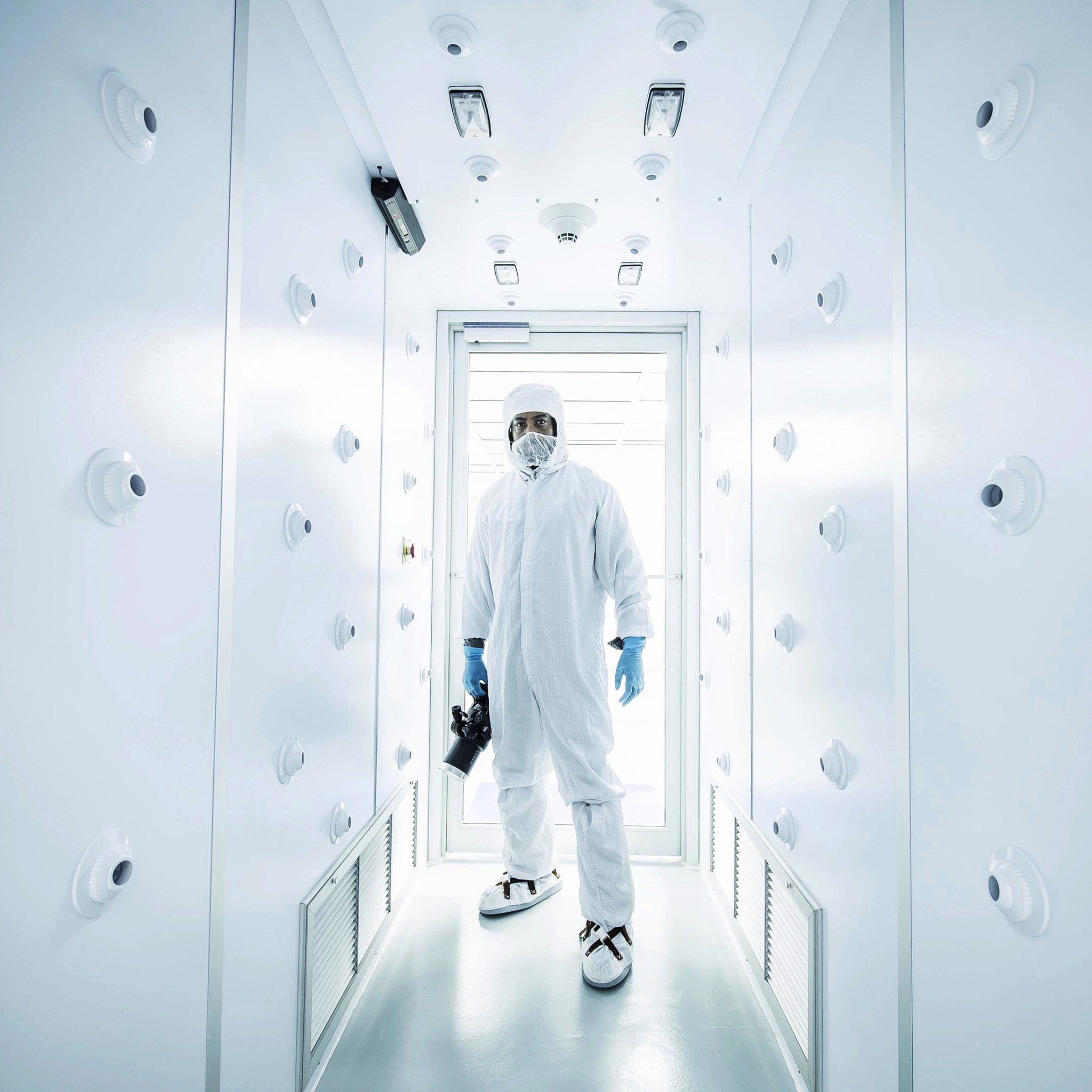 A man wearing a head-to-toe white suit and blue gloves stands in a bright white hallway holding a camera.