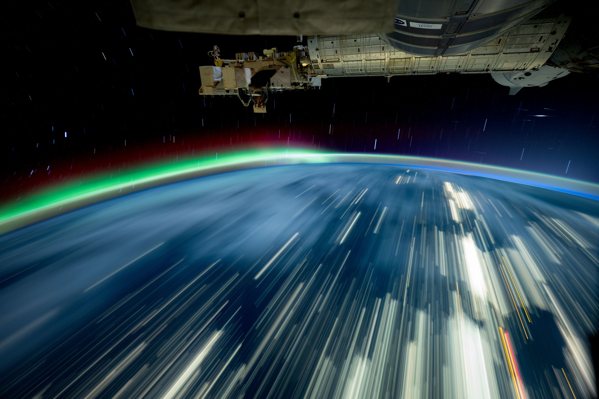 Streaks of white light cover a blue Earth, and a green and red aurora rises from the horizon. The exterior of the space station is visible in the top third of the image.