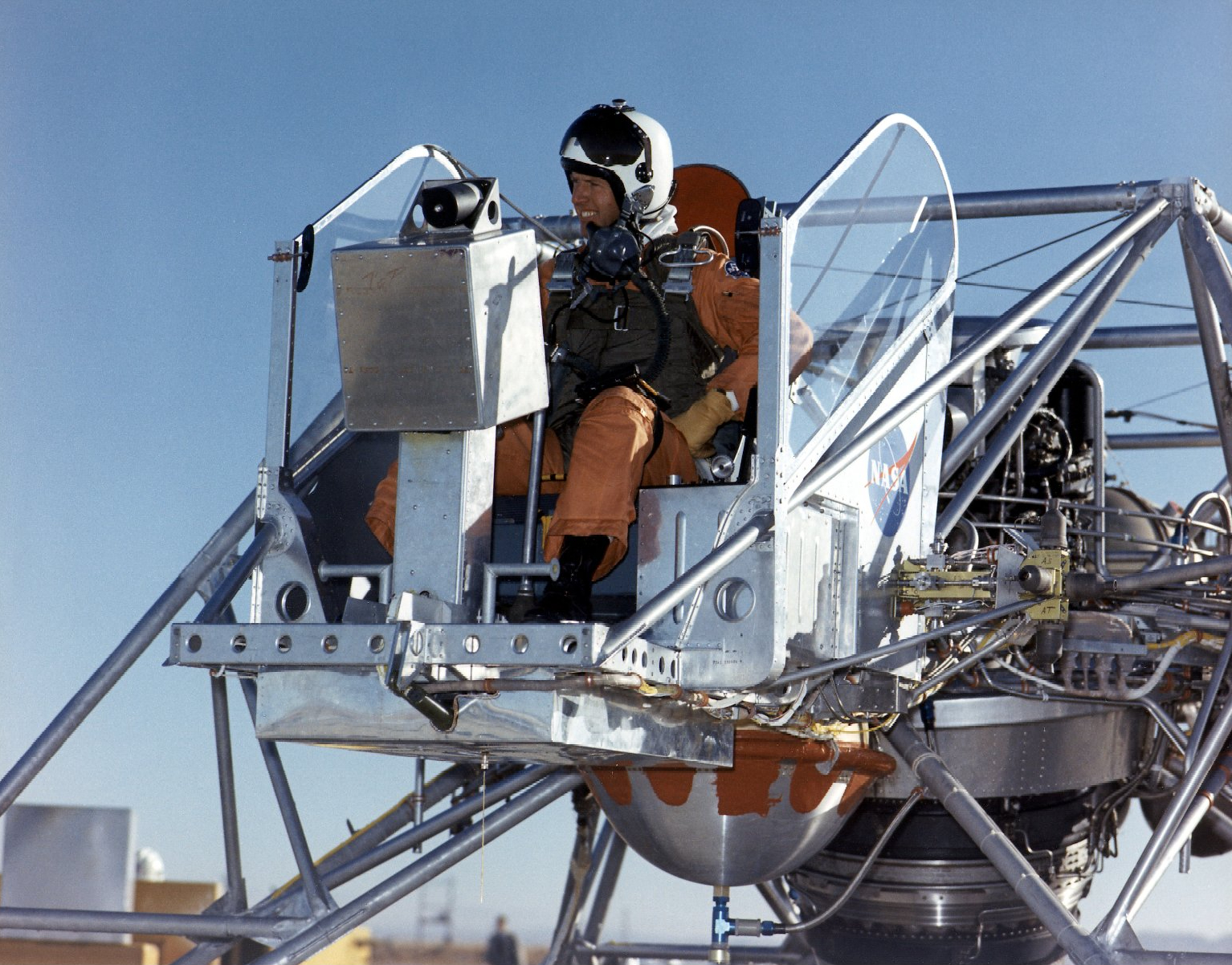 A man in an orange jumpsuit and white helmet sits in the pilot’s seat on a metal vehicle, the Lunar Landing Research Vehicle, or LLRV. The LLRV has a lot of metal tubing everywhere, which made people compare it to a metal bedframe.