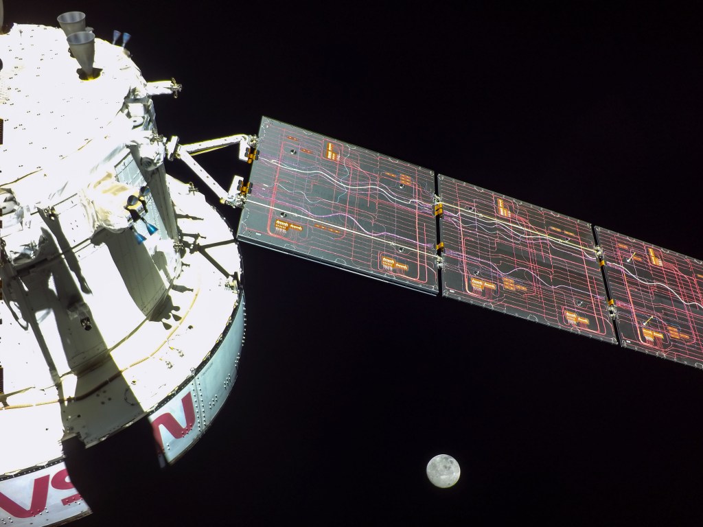 Photo of the Orion capsule, a white spacecraft with a solar array deployed, with the moon in the background.