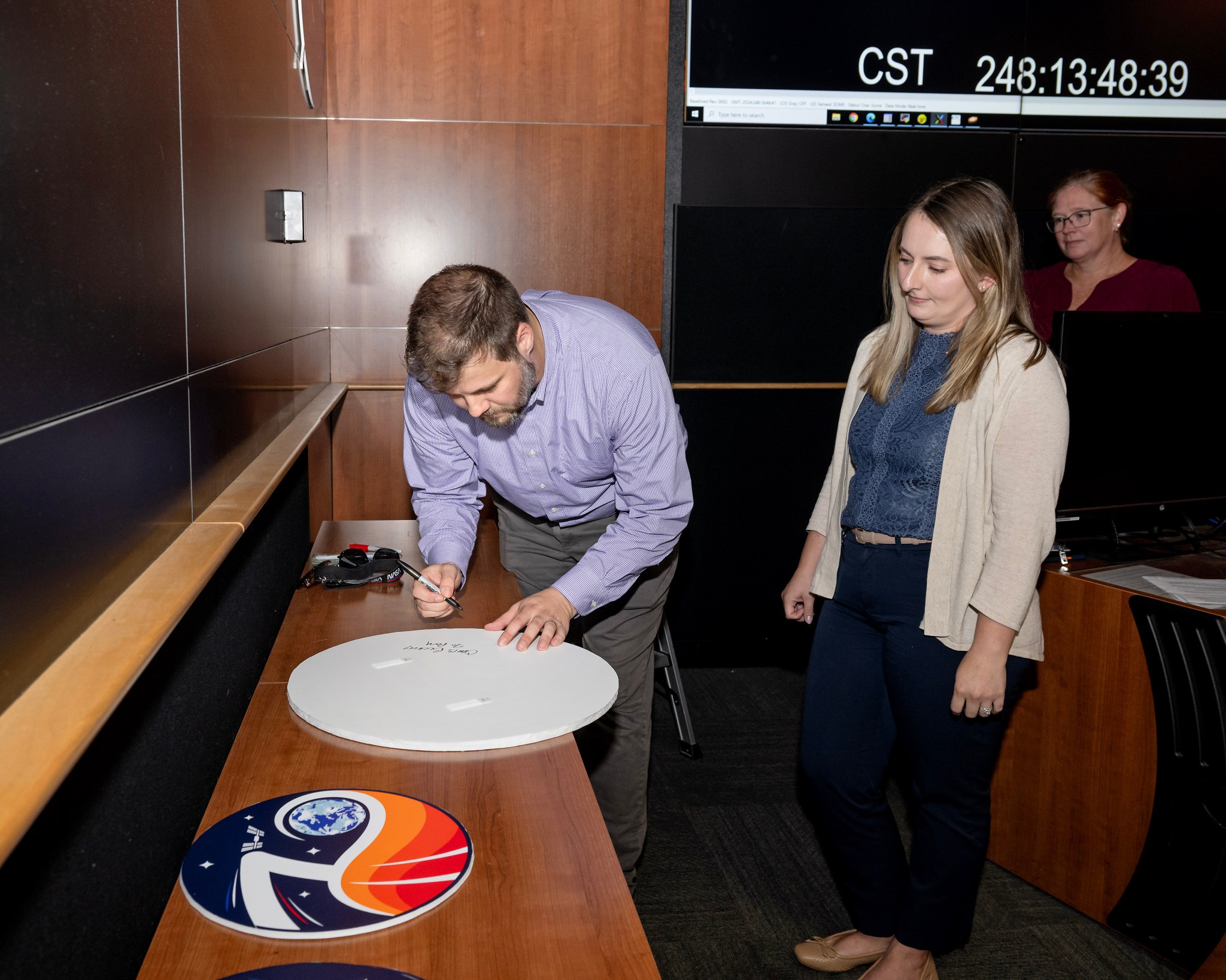 Chris Buckley, left, signs an Expedition 70 plaque as Sara Dennis, center, and Shelby Bates look on.