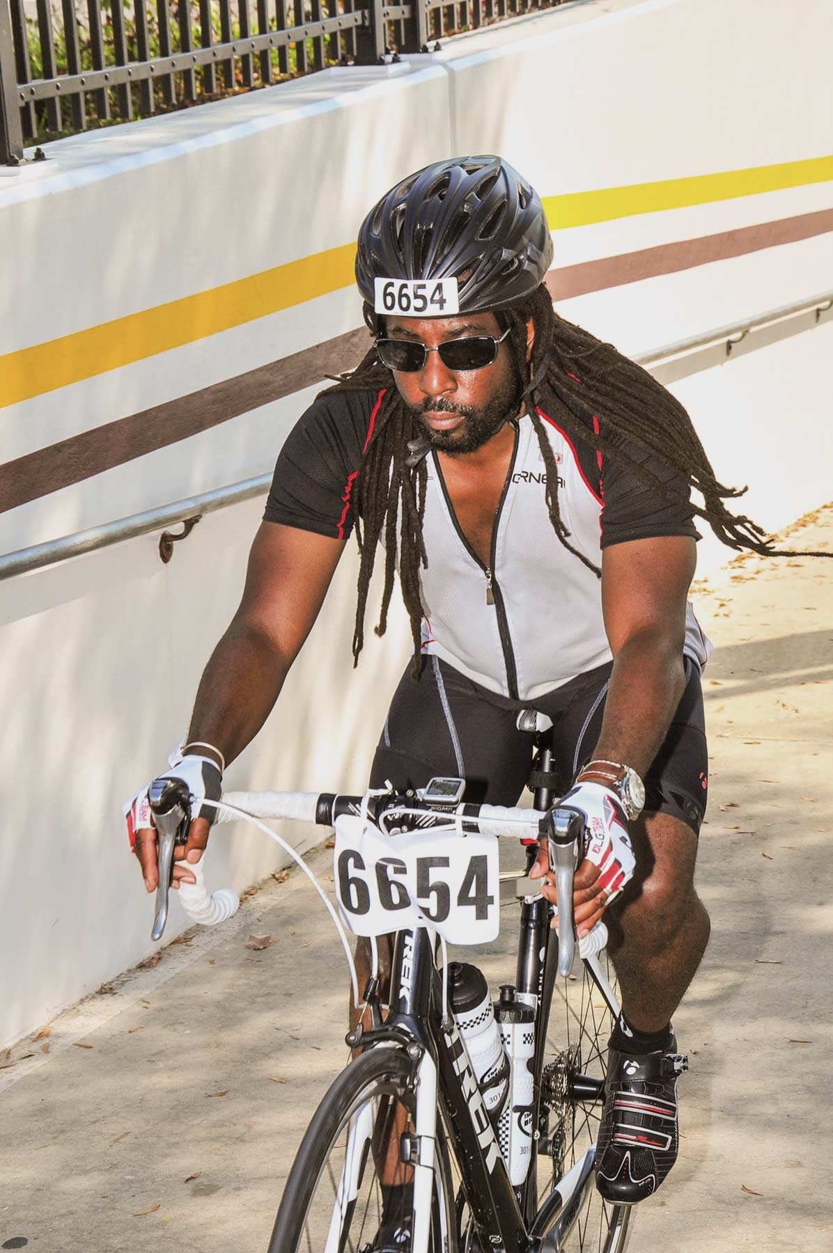 A photo of a man with long black locs riding a bike marked with the number 6654. He is wearing a black helmet marked with the same number, along with fingerless racing gloves and black sunglasses.