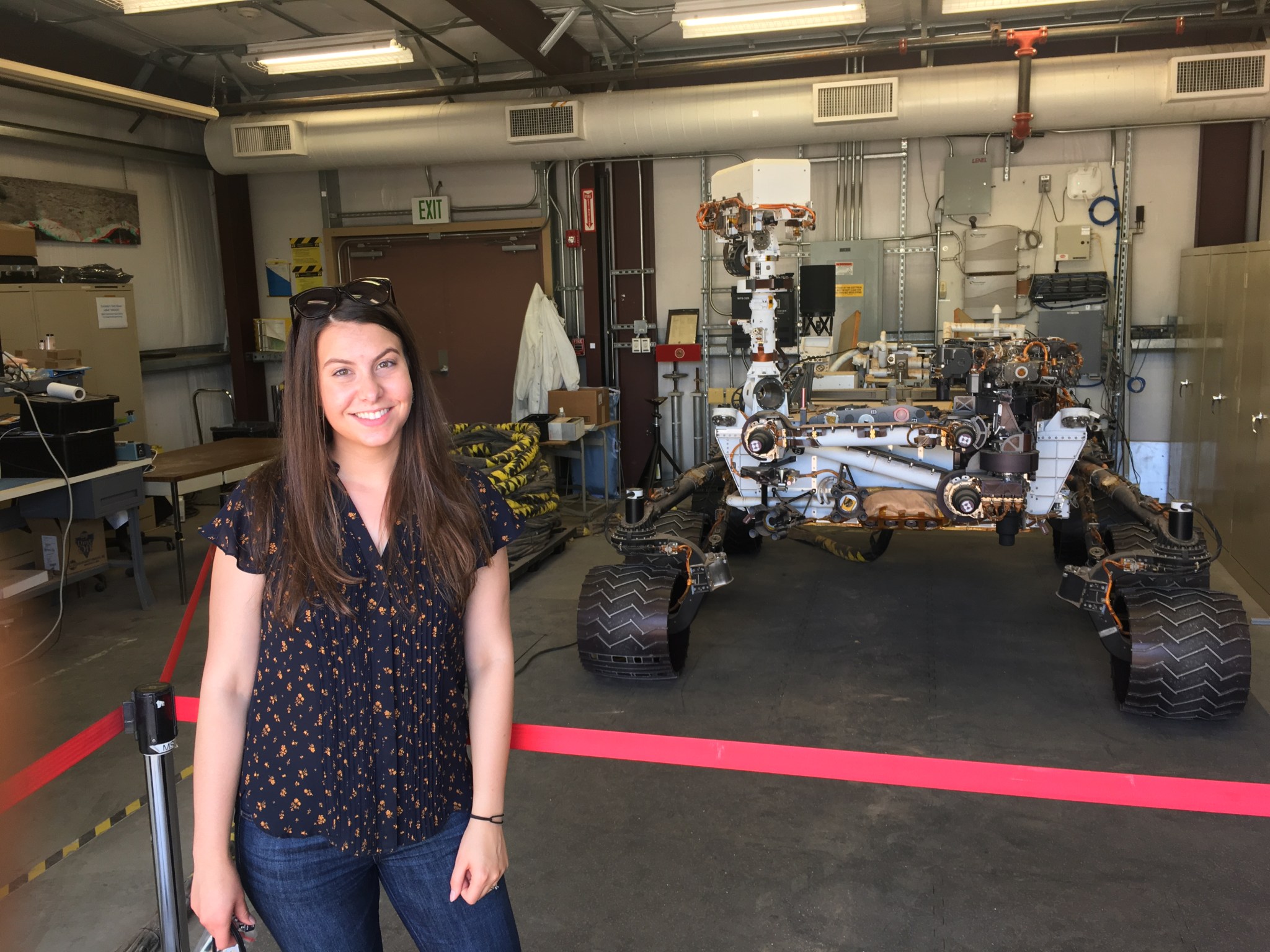 person stands in front of stanchions and red ribbon, behind which is a model of the six-wheeled, SUV-sized Curiosity rover