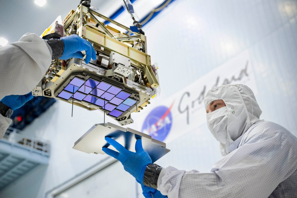 An iridescent purplish-blue array of squares attached to a chunky, rectangular piece of metal hardware is seen from below. Gloved hands guide the hardware while a man in a white suit and gloves attaches a silver covering to the array.