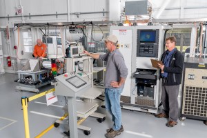 Three men work inside a building surrounded by machinery, monitors, and various equipment.