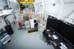 A top-down view of a large test building housing various monitors, desks, machinery, and cables. Two people sit at a desk monitoring data on computer screens while two other people operate different equipment inside the facility. 