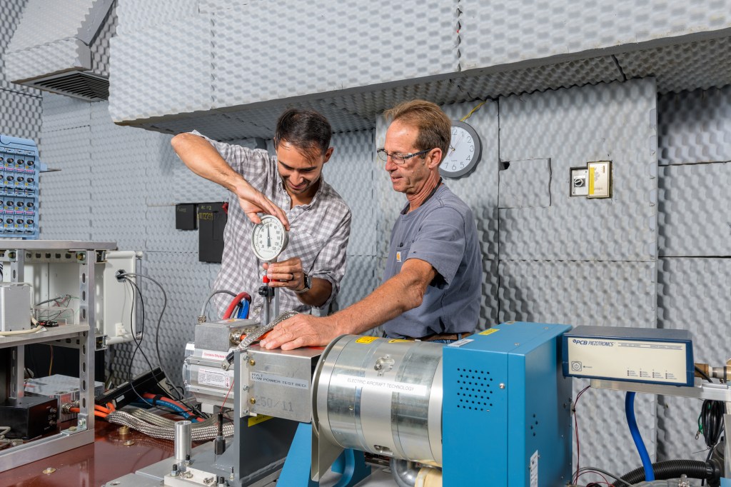 Two men adjust a device on a dynamometer surrounded by testing machinery and cables.