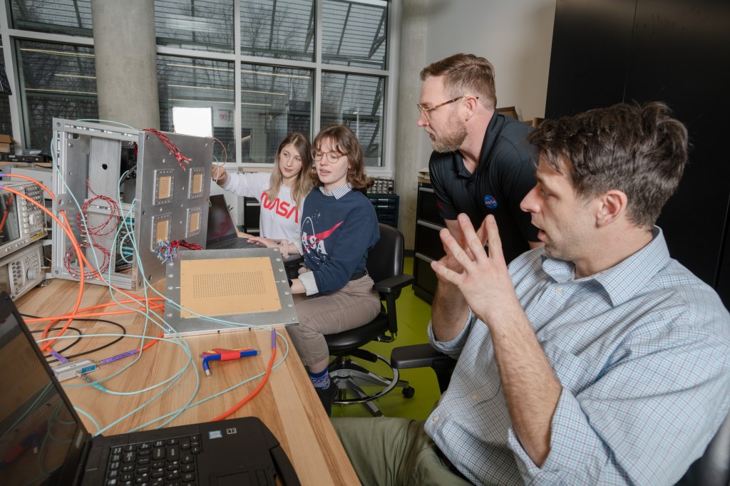 Four people sit at a table looking at a computer screen and cube shaped technology on the tabletop.