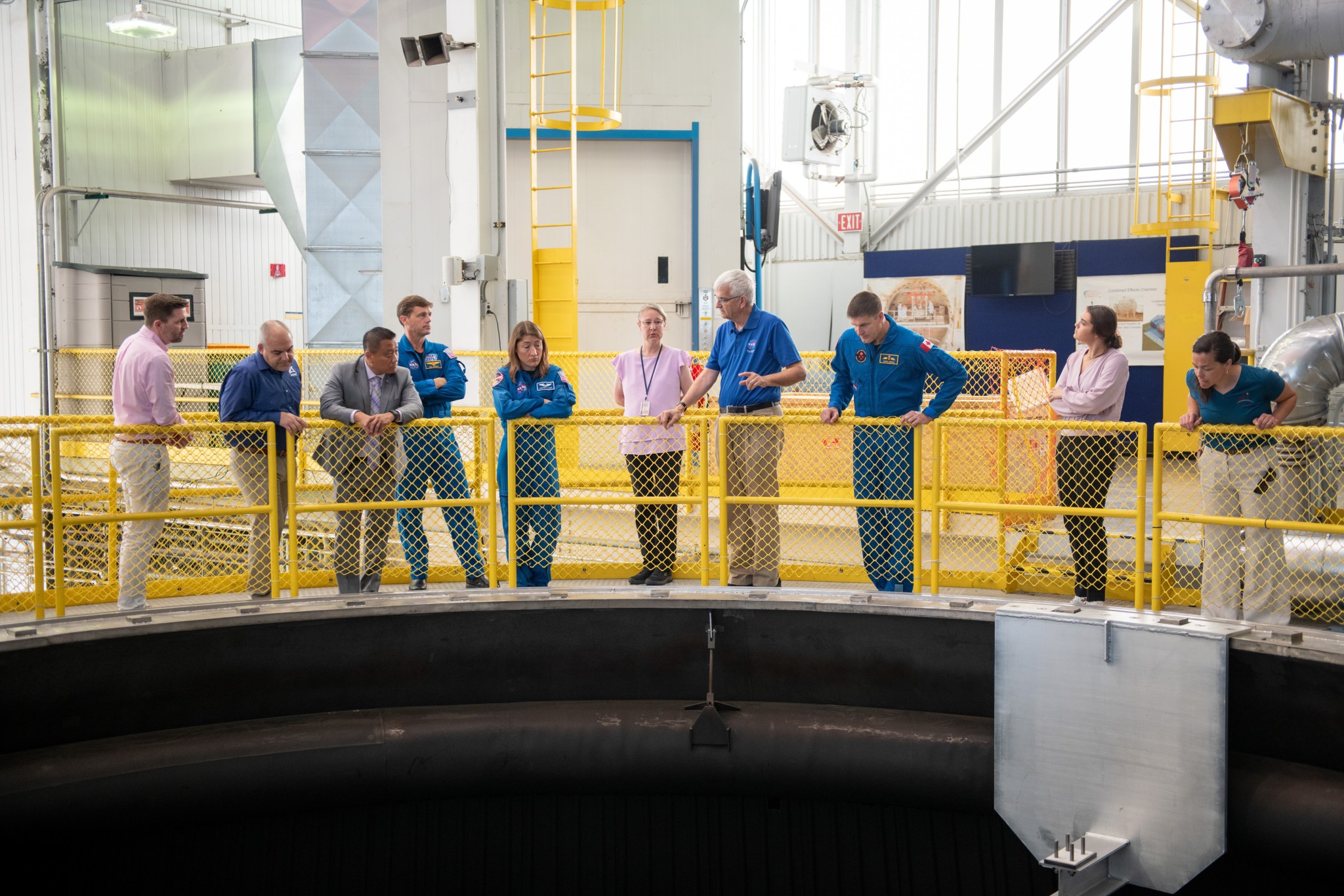 A group of people, including three astronauts wearing flight suits, stand behind yellow metal bars atop a large vacuum chamber.