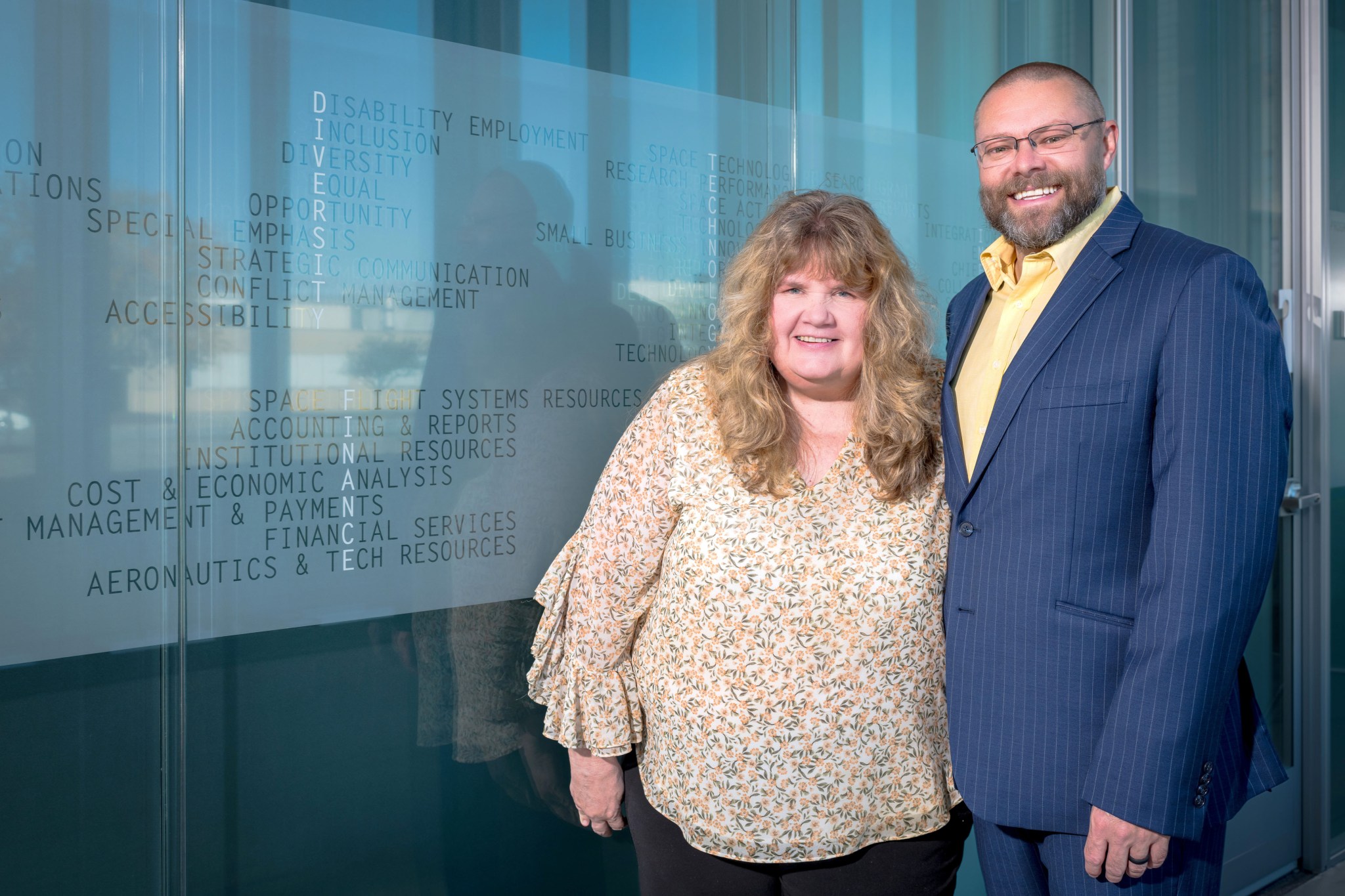 A man wearing a blue suit, a yellow shirt, and glasses and a woman wearing a yellow blouse smile at the camera as they pose standing side by side in front of a glazed glass wall. Small words are etched on the wall, including “Disability Employment, Inclusion, Diversity, Equal Opportunity, Accessibility,” and more.