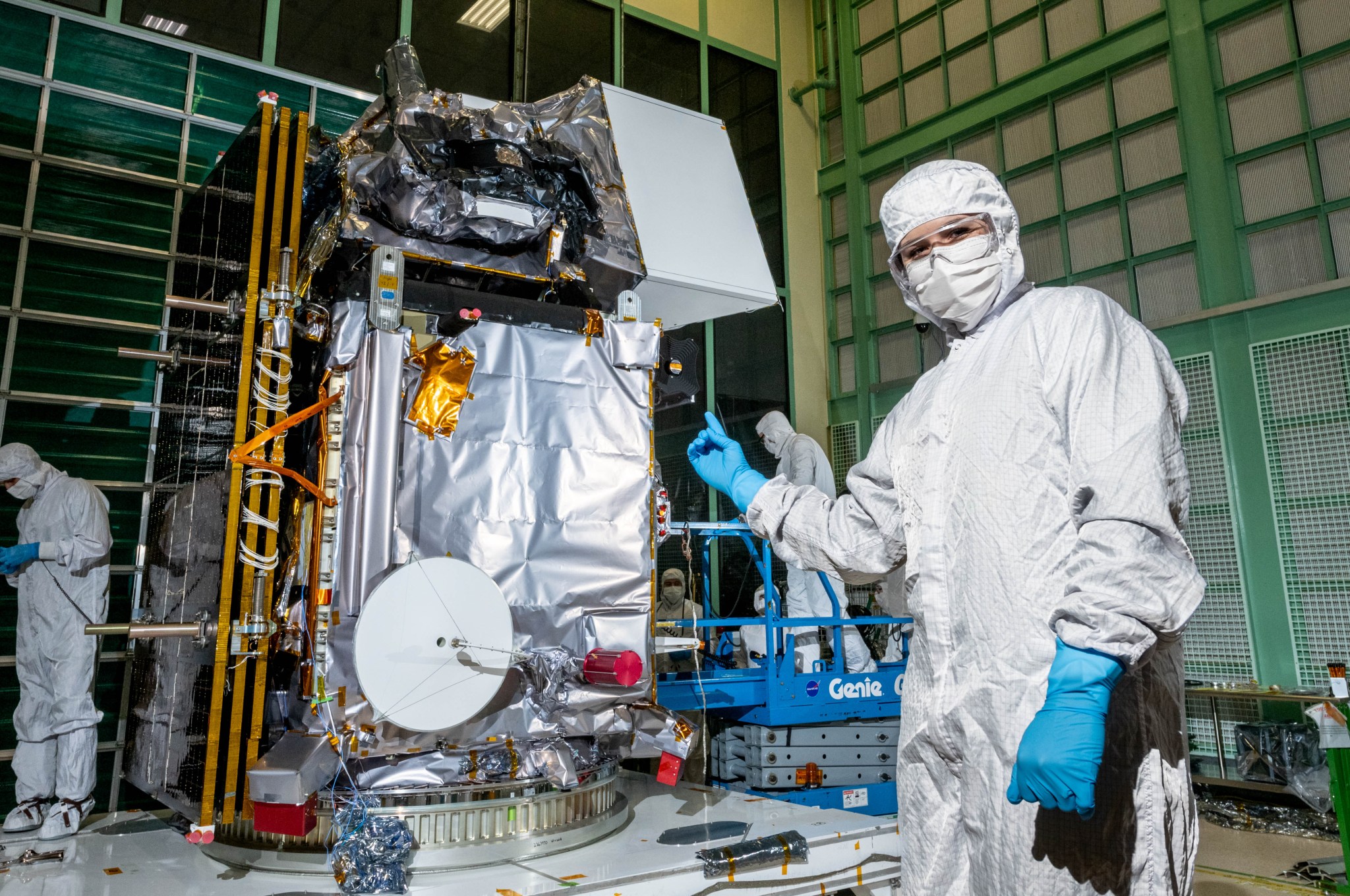 Carissa Arillo stands to the right of the PACE observatory in a cleanroom and points at it. She is wearing a white clean room suit that covers her torso, arms, and over the top of her head and forehead. She also wears a white mask that covers her nose and mouth and blue latex gloves. Behind her and slightly to the left is the PACE observatory, which is large, silver colored, and is covered in wires and other metallic pieces of instrumentation and materials. 