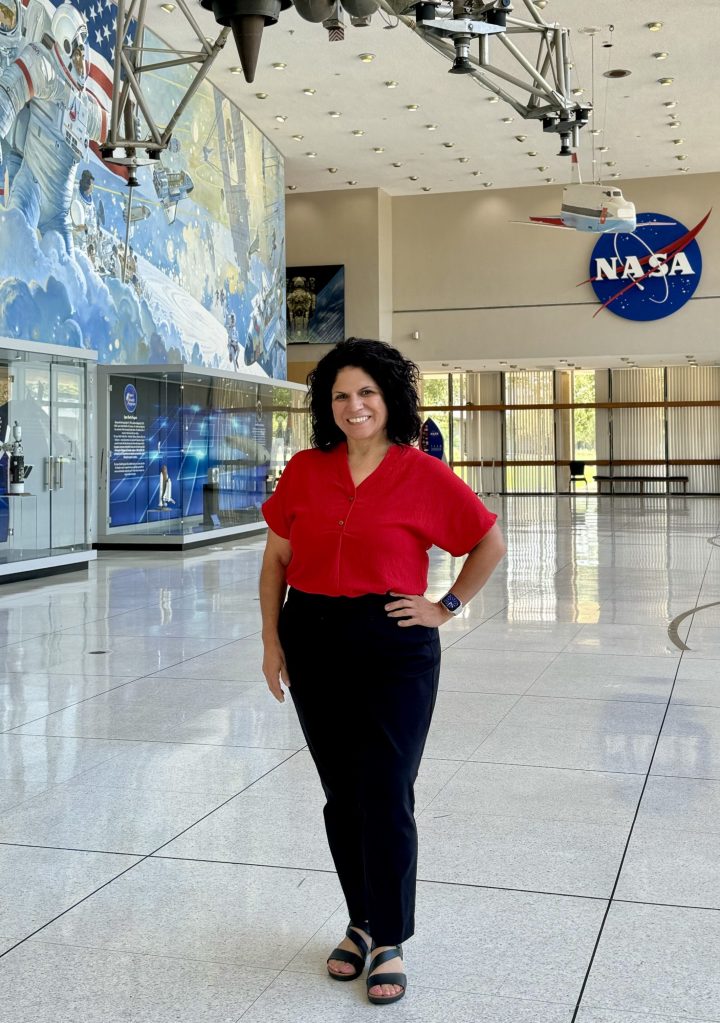 A woman stands and smiles in the Teague lobby at NASA Johnson. She is wearing a red blouse and black pants. Behind her is a mural of astronauts and space exploration, as well as a NASA logo on the wall.