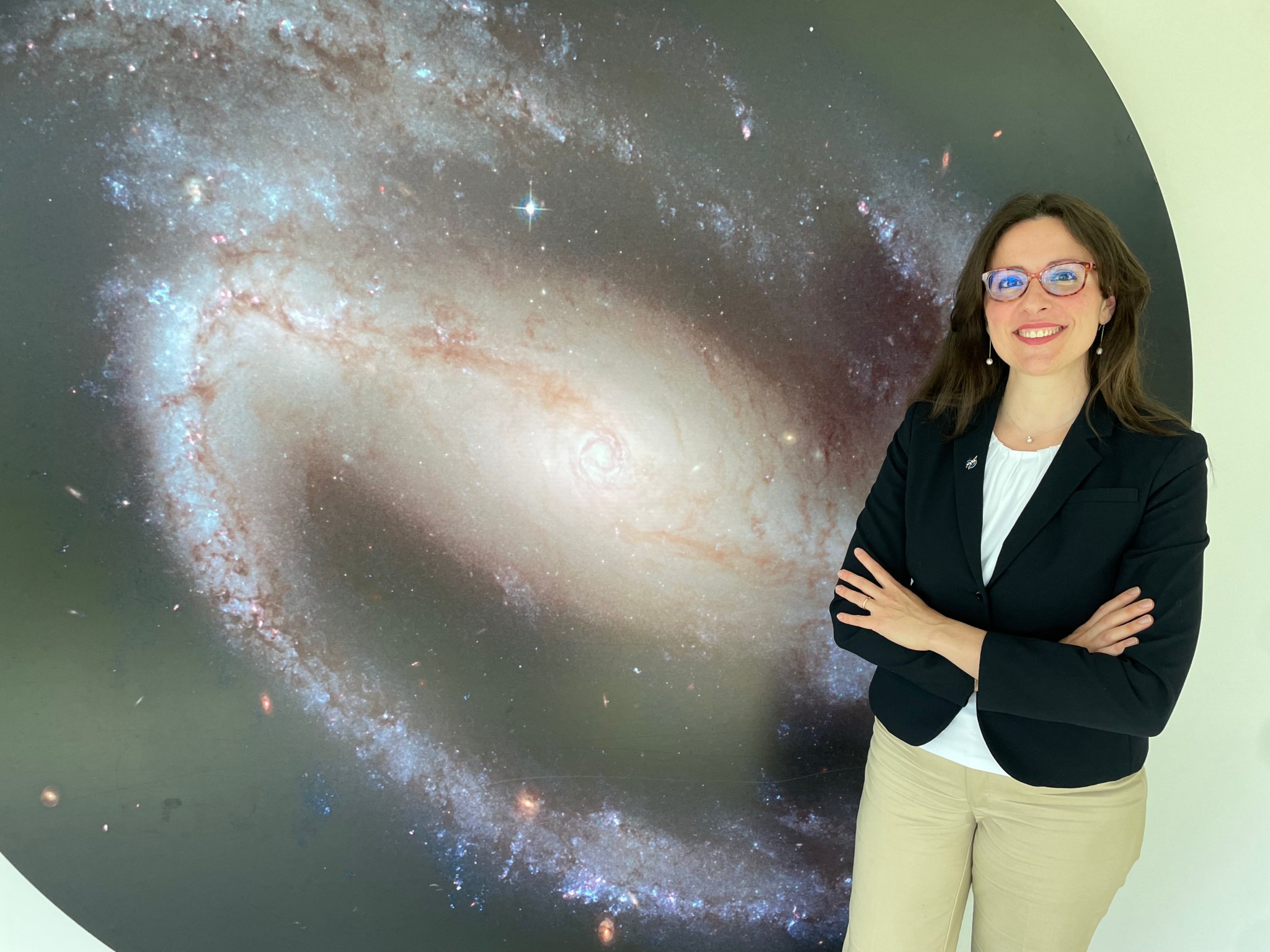 Dr. Gioia Rau stands against a wall with a large Hubble image of a spiral galaxy. She is standing to the right of the image wearing a black blazer, white shirt, tan pants, and glasses. 