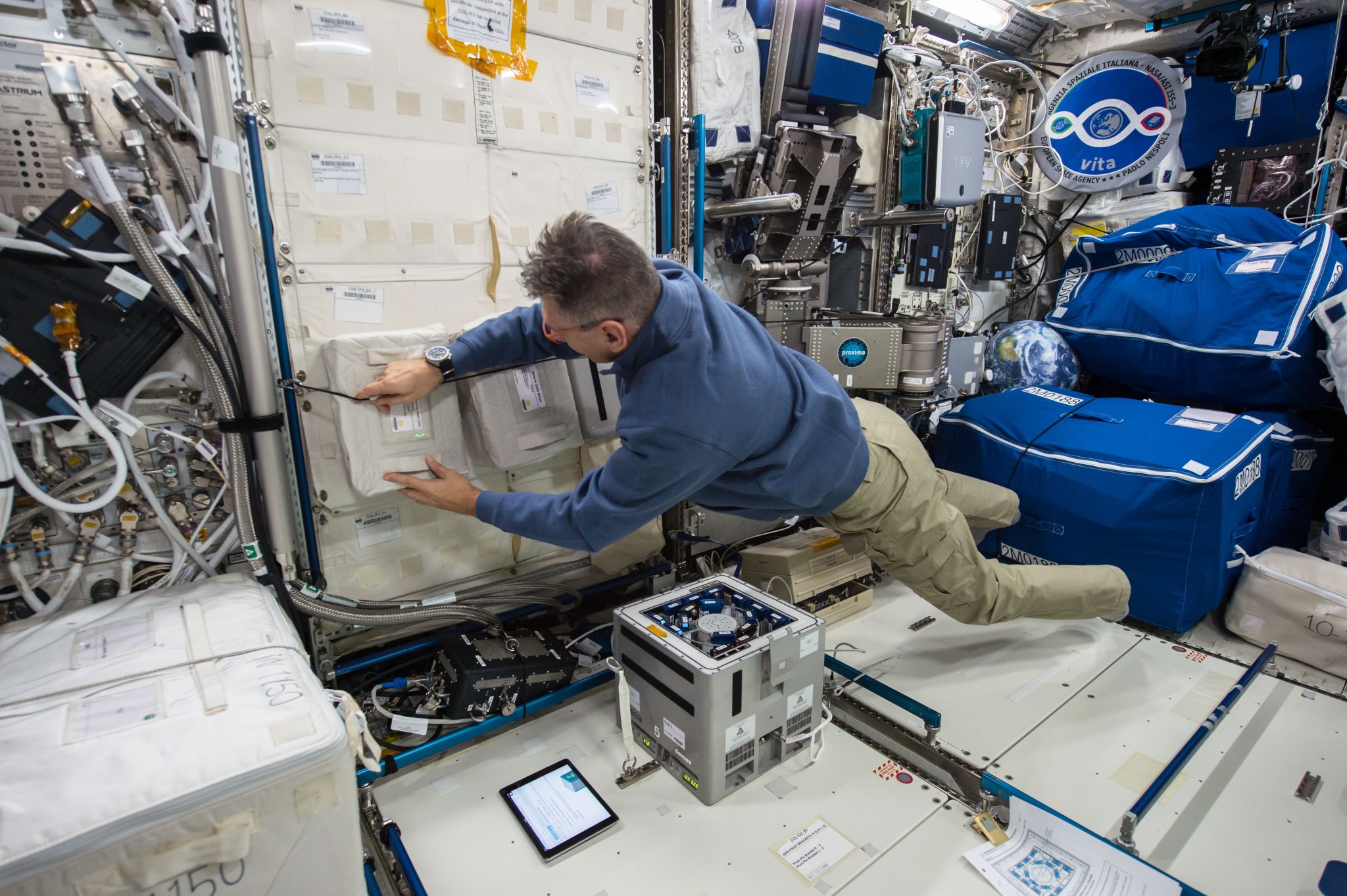 NASA astronaut working inside the International Space Station, floating in a microgravity environment while securing a storage container to the wall.