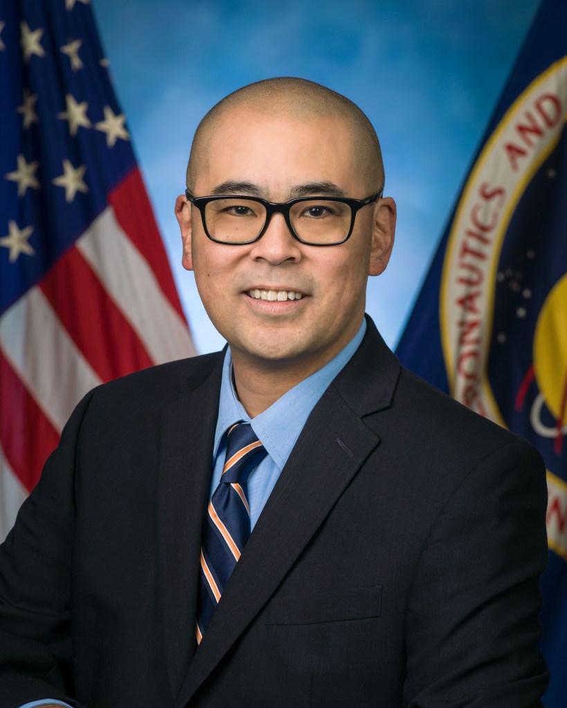 A professional headshot of a man wearing glasses, a dark suit, and a striped tie, standing in front of the American flag and a NASA emblem.