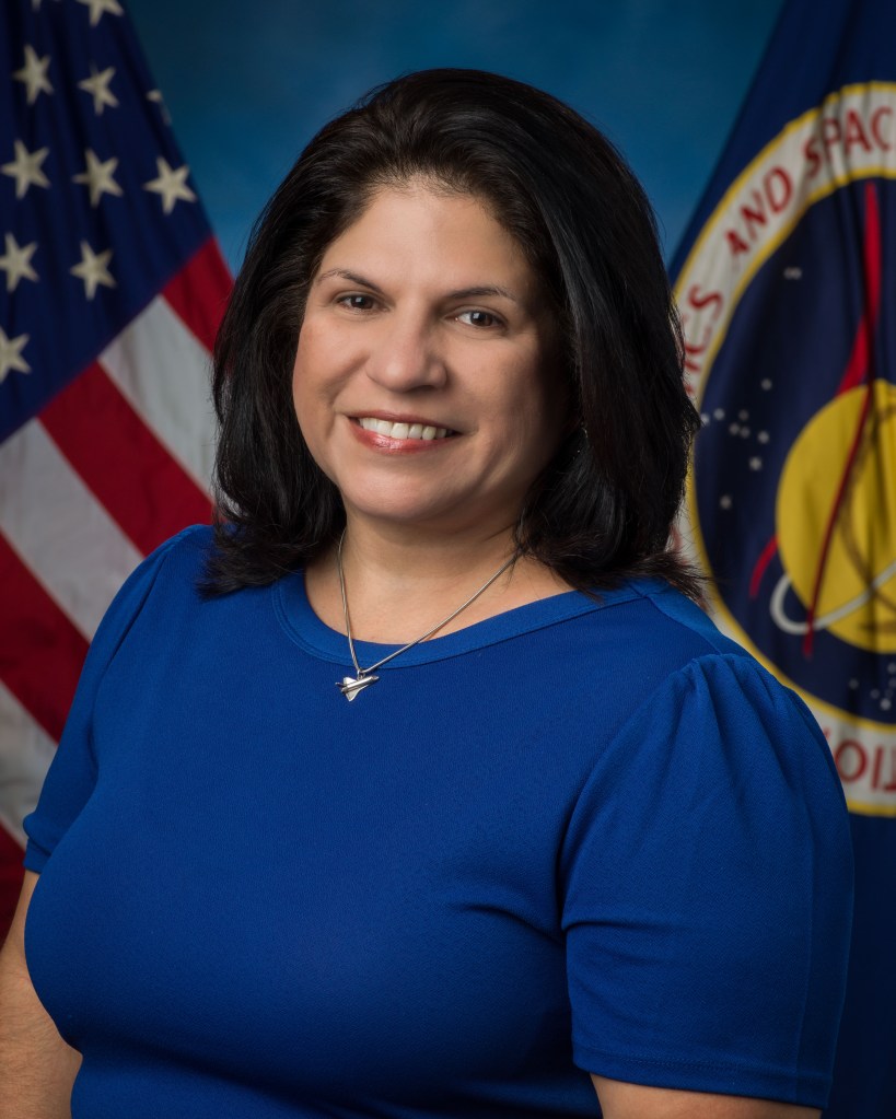 A professional portrait of a woman with dark hair, wearing a blue top. She stands in front of the American flag and NASA’s emblem.