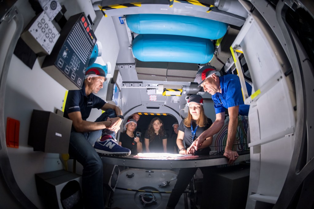 Engineers and astronauts gather inside the mock-up of the ESA Lunar I-Hab module during a 'human in the loop' test. The group, wearing protective caps, discusses the module's interior layout while reviewing design plans under a well-lit table. Mock-up equipment and placeholders surround the participants, simulating the environment of the habitation module that will eventually be part of Gateway.