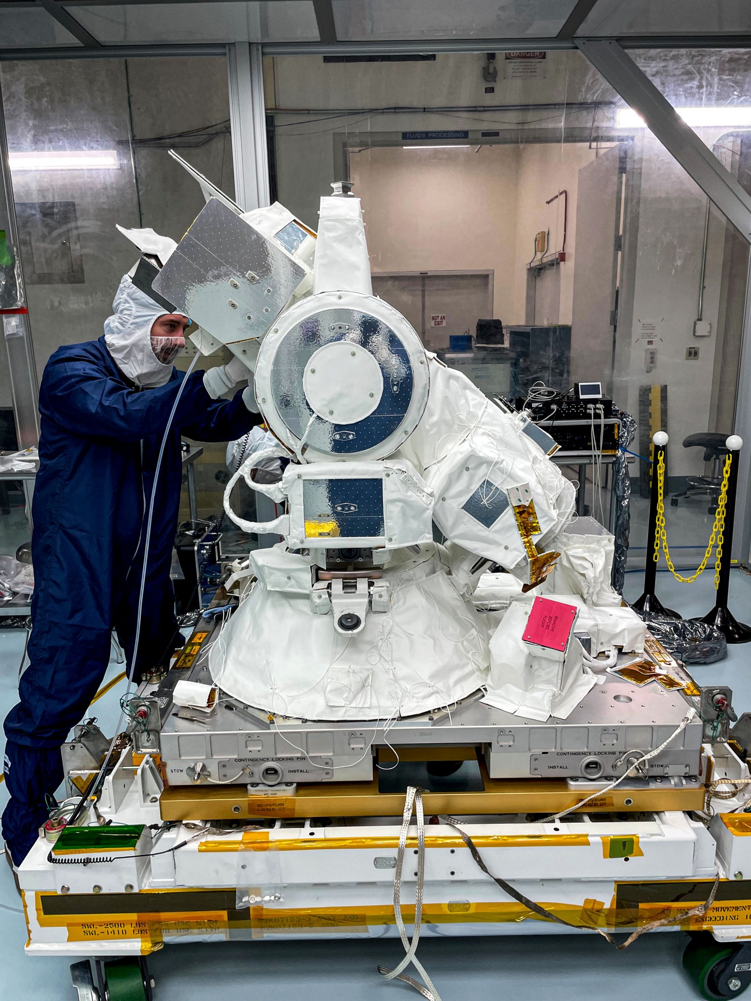 A person in a blue clean suit and white hood is visible to the left of and a little behind a large instrument that looks like a telescope, with a wide base and a large arm at an angle on top of it. The instrument has several silver panels, and much of it is covered in white protective fabric.