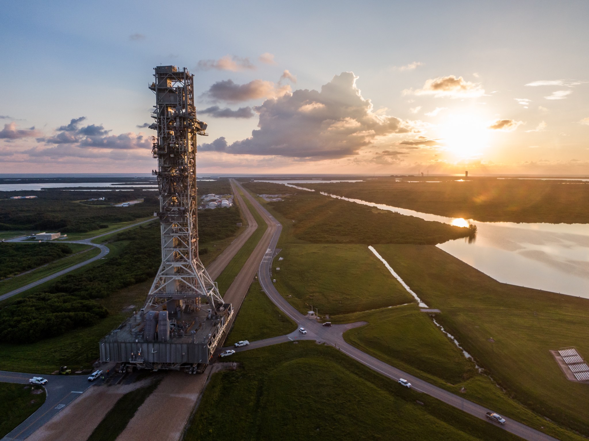 A large rectangular structure - the mobile launcher - rests atop a moving rectangular base - the crawler-transporter. In this aerial shot, they are moving along a road. Green grass covers the surrounding area.