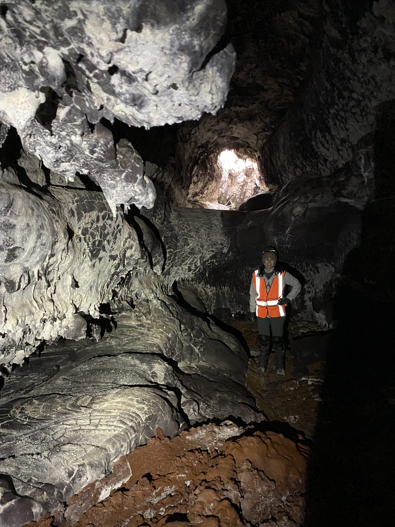 a person wearing an orange reflective safety vest stands in a dark gray, rocky lava tube tunnel