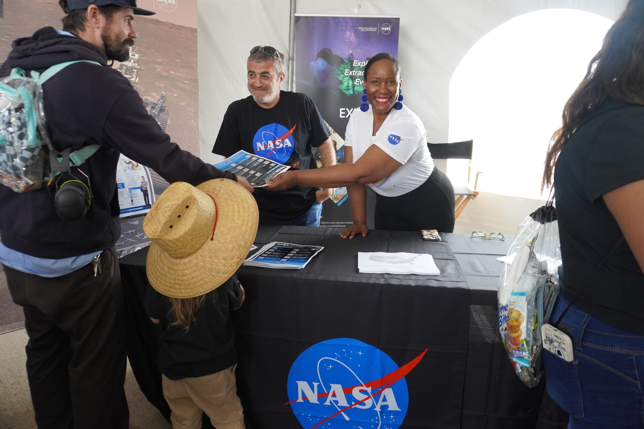 A woman smiles at the camera as she hands a guest and child a flier from behind a NASA table. The guest receiving the flier smiles back at her and a small child in a straw hat stands beside him. Next to the woman is a man smiling at guests behind the patron at the table. The NASA meatball sign is visible on the man’s tee shirt, the woman’s sleeve, and the tablecloth. Behind them, posters of airborne NASA missions are partially visible.