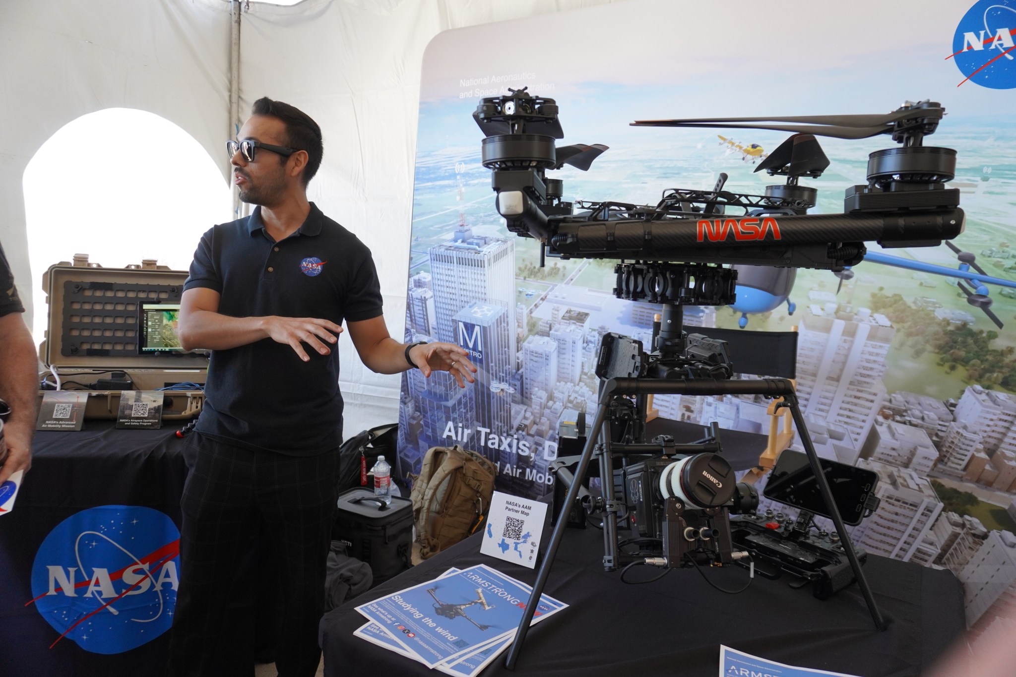 A man stands beside a table featuring a drone exhibit. He seems to be in the middle of explaining the drone to an unseen guest, using his hands to gesture toward the drone. He is wearing a navy polo with a NASA meatball on the chest and sunglasses. Behind him is a partially visible science instrument featured on top of a neighboring exhibit table. Behind him and the tables is a large poster of NASA’s Advanced Air Mobility program.