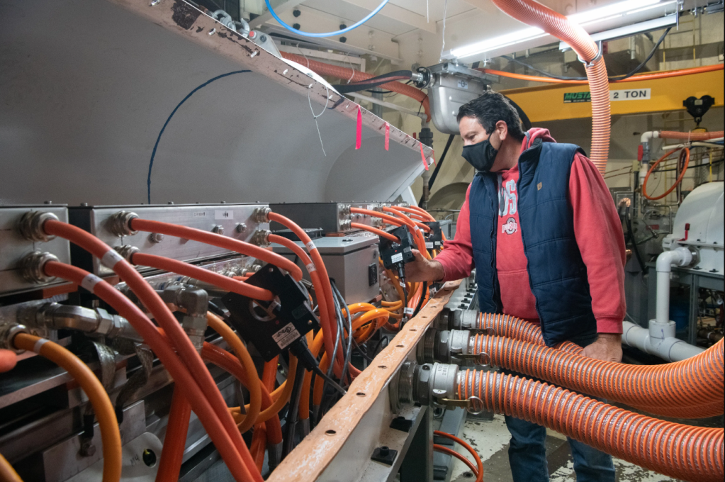 A man wearing a mask inspects electrified aircraft propulsion components inside a test rig filled with various equipment and orange cables.