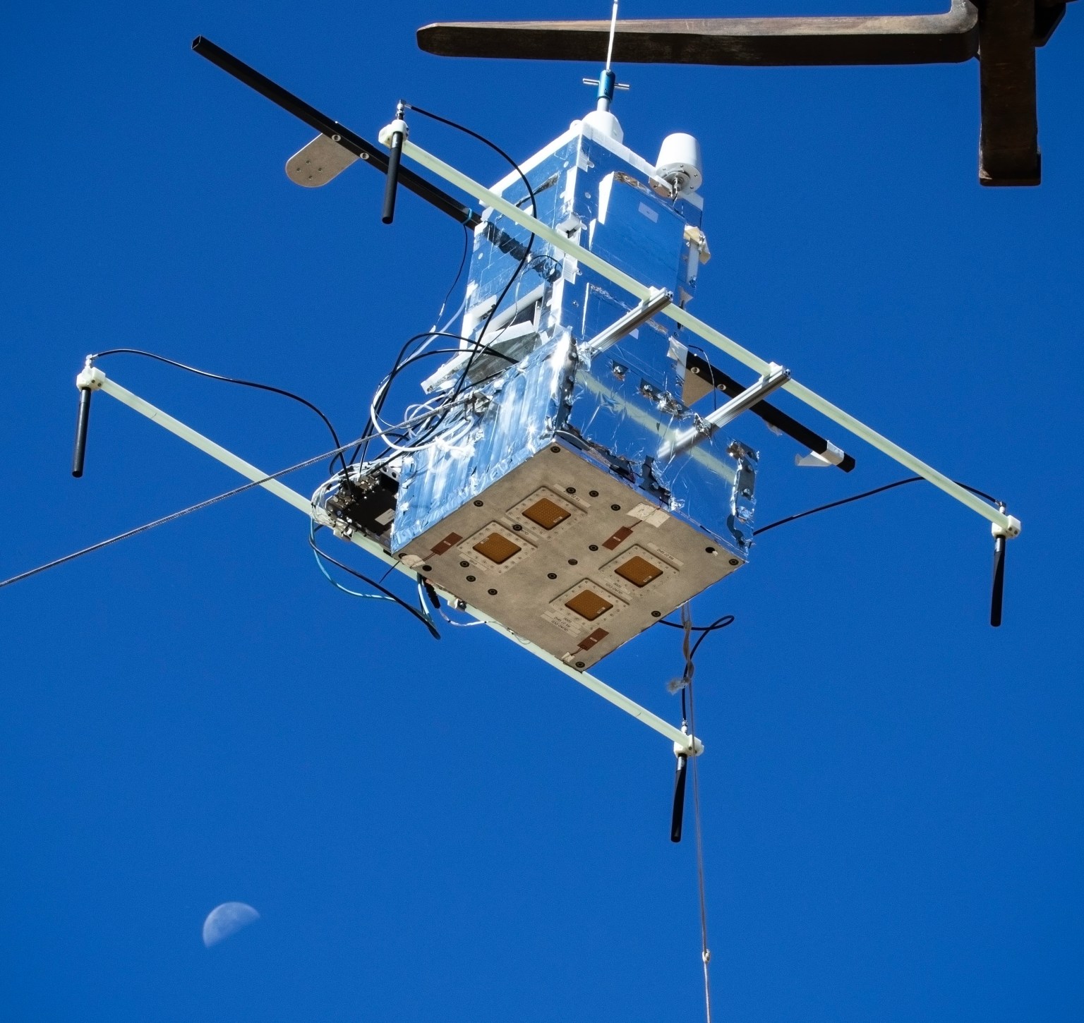 View from below of a metal cube with four inset squares suspended by a frame against a blue sky.