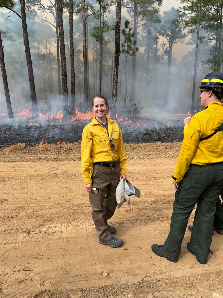 A white woman in a yellow long-sleeve shirt and khaki fire pants smiles at the camera while standing on tan dirt. In the background a fire burns the ground of a forest.