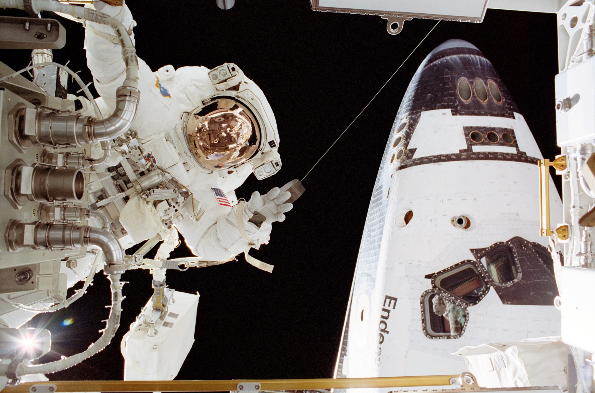 An astronaut in a white spacesuit appears on the left side of the image near a part. On the right in  the back is part of the space shuttle Endeavour.