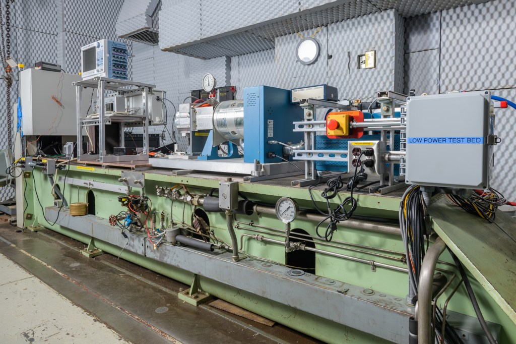 An angled view of a large, long testbed setup featuring metal hardware, wires, cables, and testing machinery.