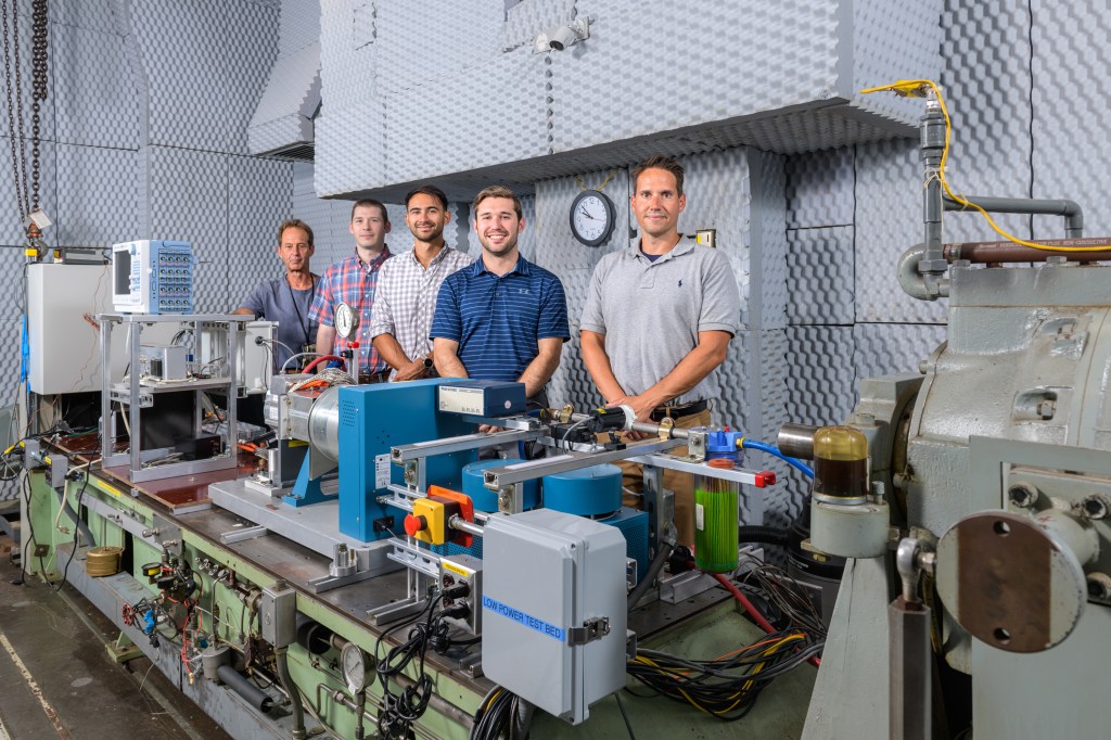 A group of five men pose behind a testing rig made up of various hardware, wires, and testing equipment.