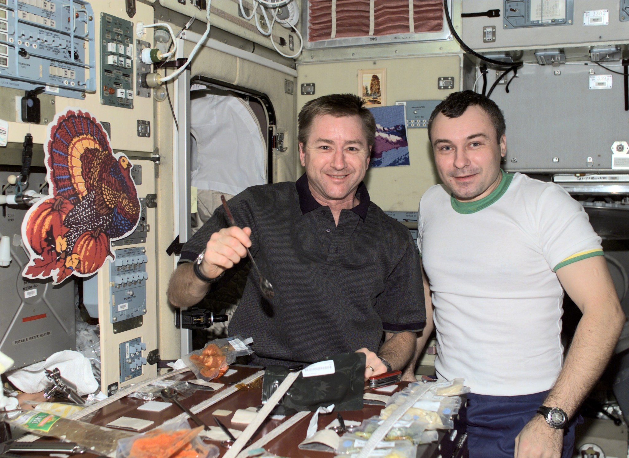 A man in a black shirt is on the left and a man in a white shirt is on the right. Both are eating Thanksgiving dinner together on a spacecraft. They are surrounded by equipment. The astronaut in the black shirt on the left is holding a spoon in his right hand.