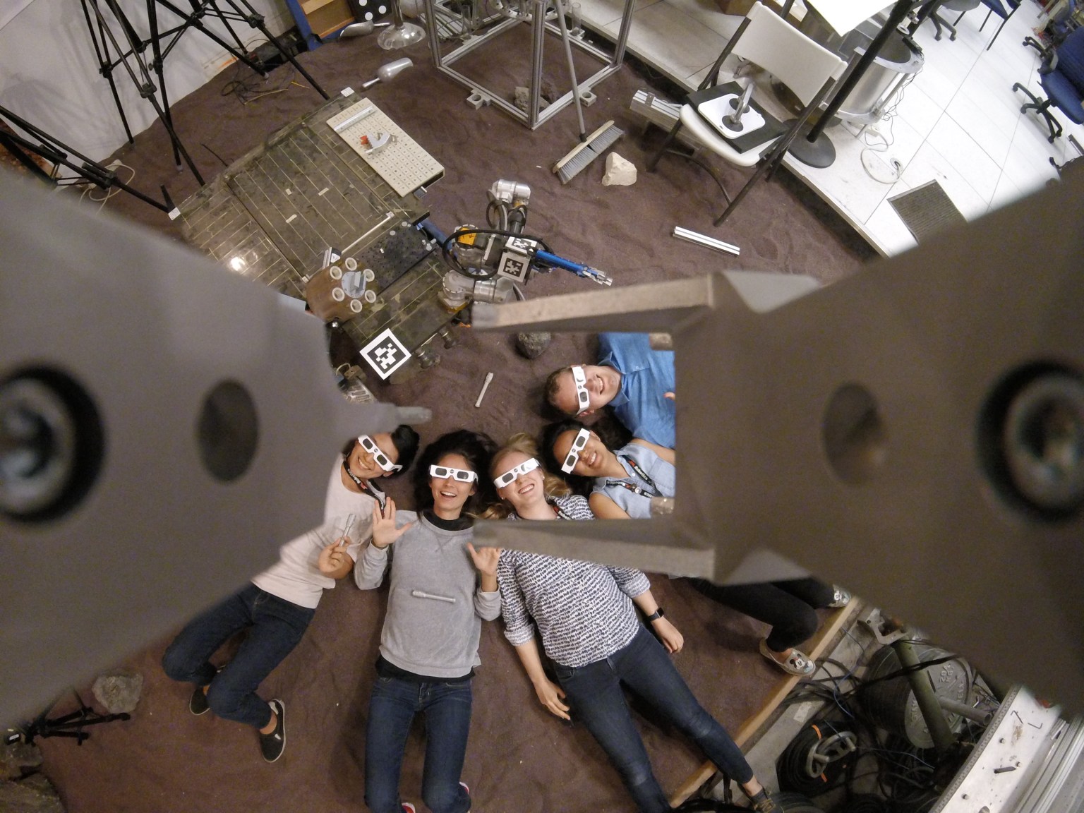 A group of male and female interns at NASA's Jet Propulsion Laboratory pose for photos in eclipse glasses. Looking up in a circle the group smiles and wears business casual attire laboratory instruments and devices are visible around the peripheral of the image. Credit: NASA/JPL-Caltech