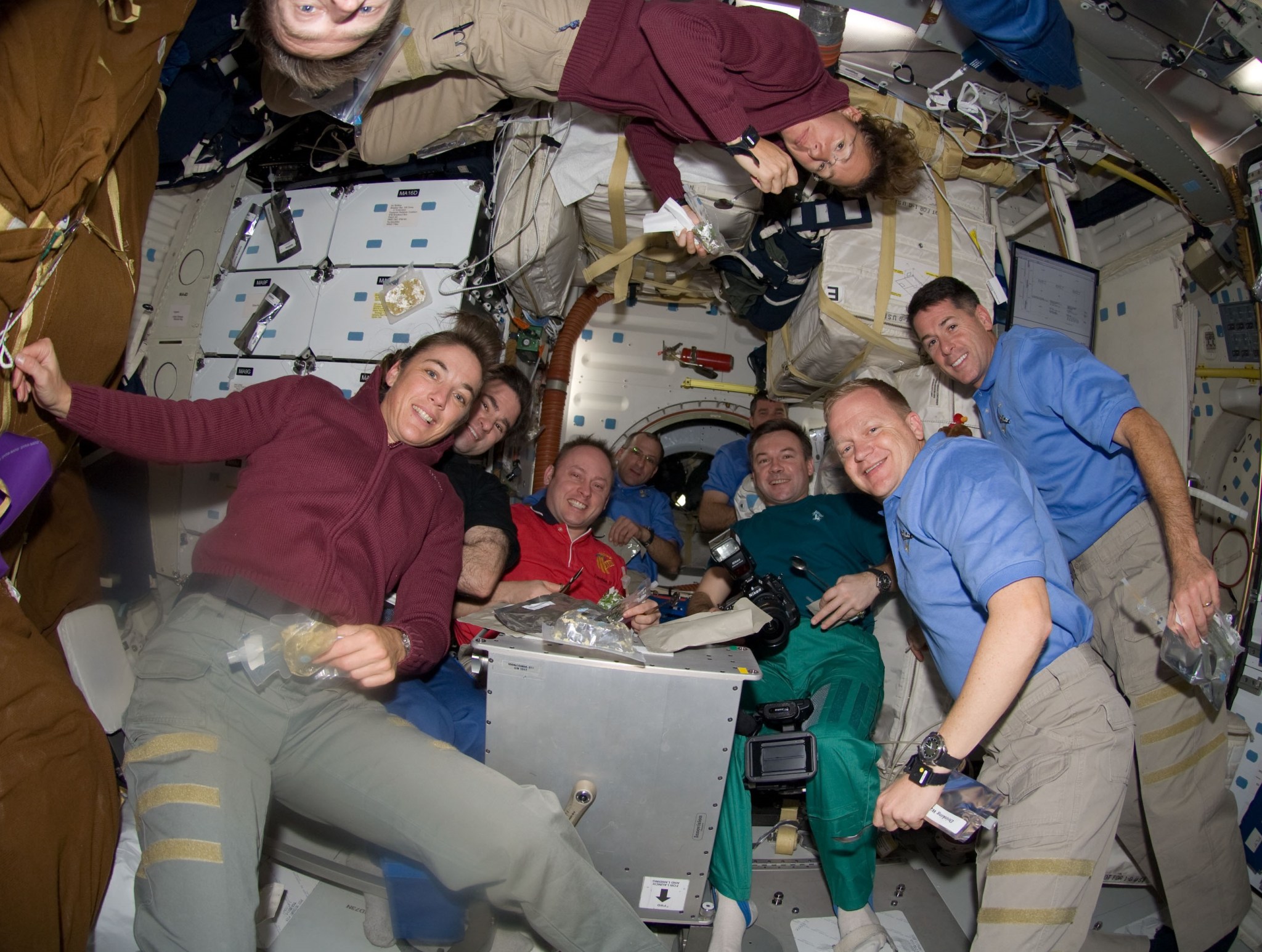 Nine astronauts are sharing a meal together while floating middeck of on NASA's space shuttle. Three of the astronauts are wearing red shirts and five of them are wearing blue. One astronaut is wearing a black shirt. One astronaut in a red shirt is floating upside down. 