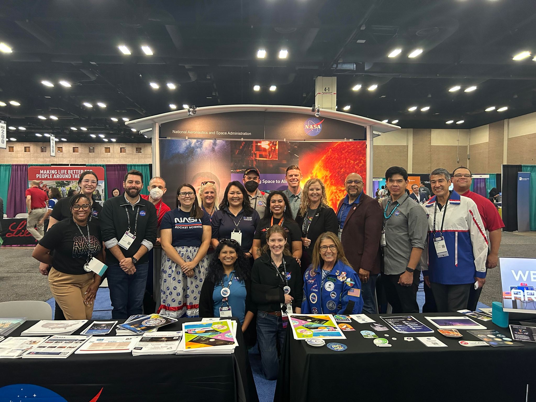 A group of 18 NASA employees pose in front of tables filled with various flyers, fact sheets, and handouts. Behind them is a large NASA backdrop with images of the Sun and deep space, as well as other conference booths.