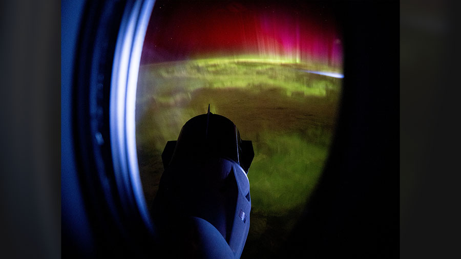 The SpaceX Dragon Freedom spacecraft is pictured through the window of the SpaceX Dragon Endeavour spacecraft with a vivid green and pink aurora below.