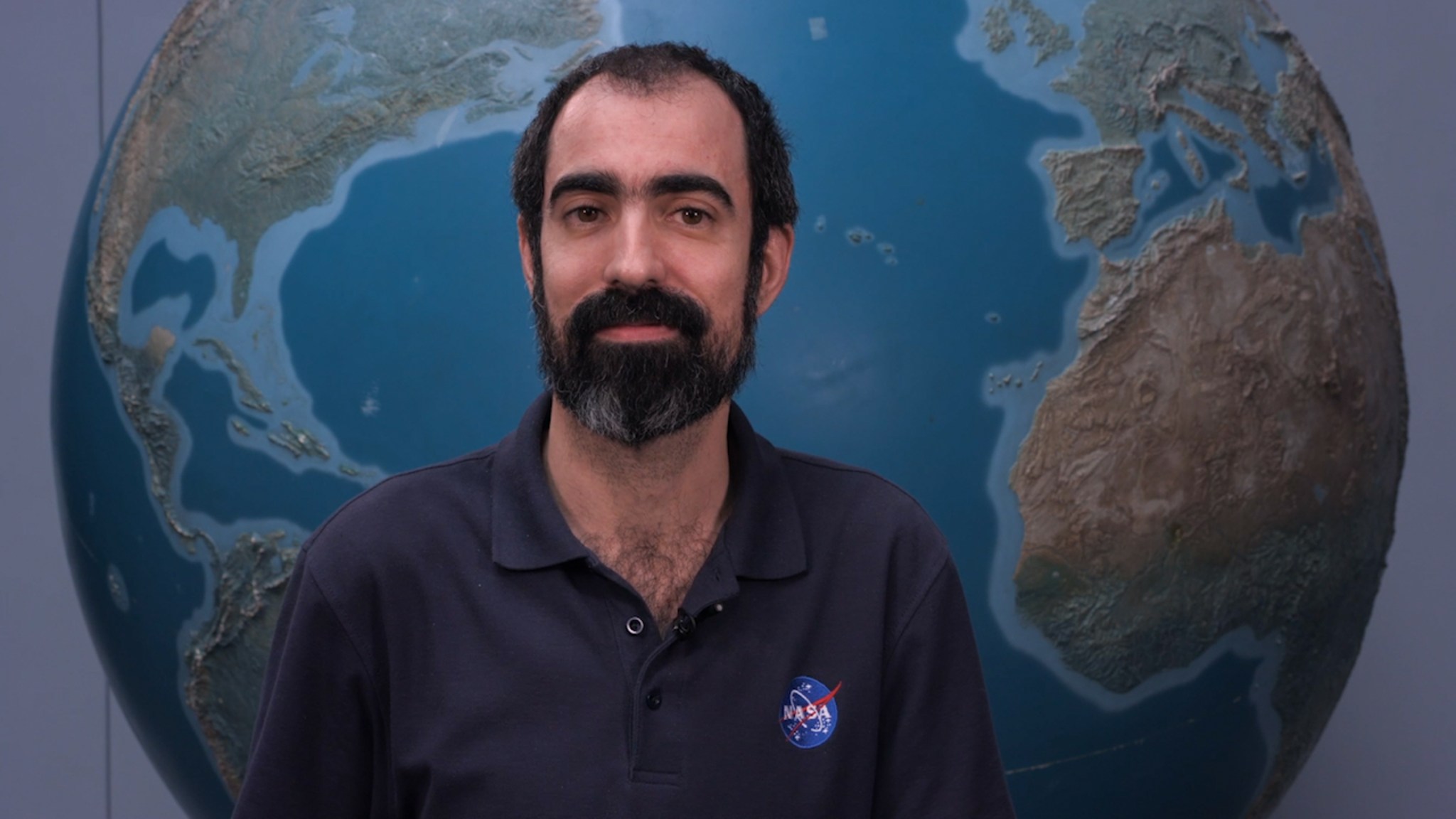 Alfonso stands in front of a large globe of Earth. He has dark hair and a beard and mustache, with a thoughtful expression. He is wearing a navy blue polo with the blue, red, and white NASA “meatball” logo on the chest patch.