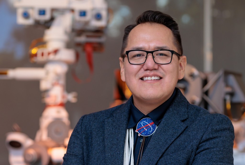 Aaron Yazzie, a Navajo man with black hair and glasses, stands with his arms crossed. He wears business professional attire with a navy blue jacket and beaded Indigenous tie making the NASA meatball logo. Behind him a large model of NASA's Mars Perseverance rover is visible.