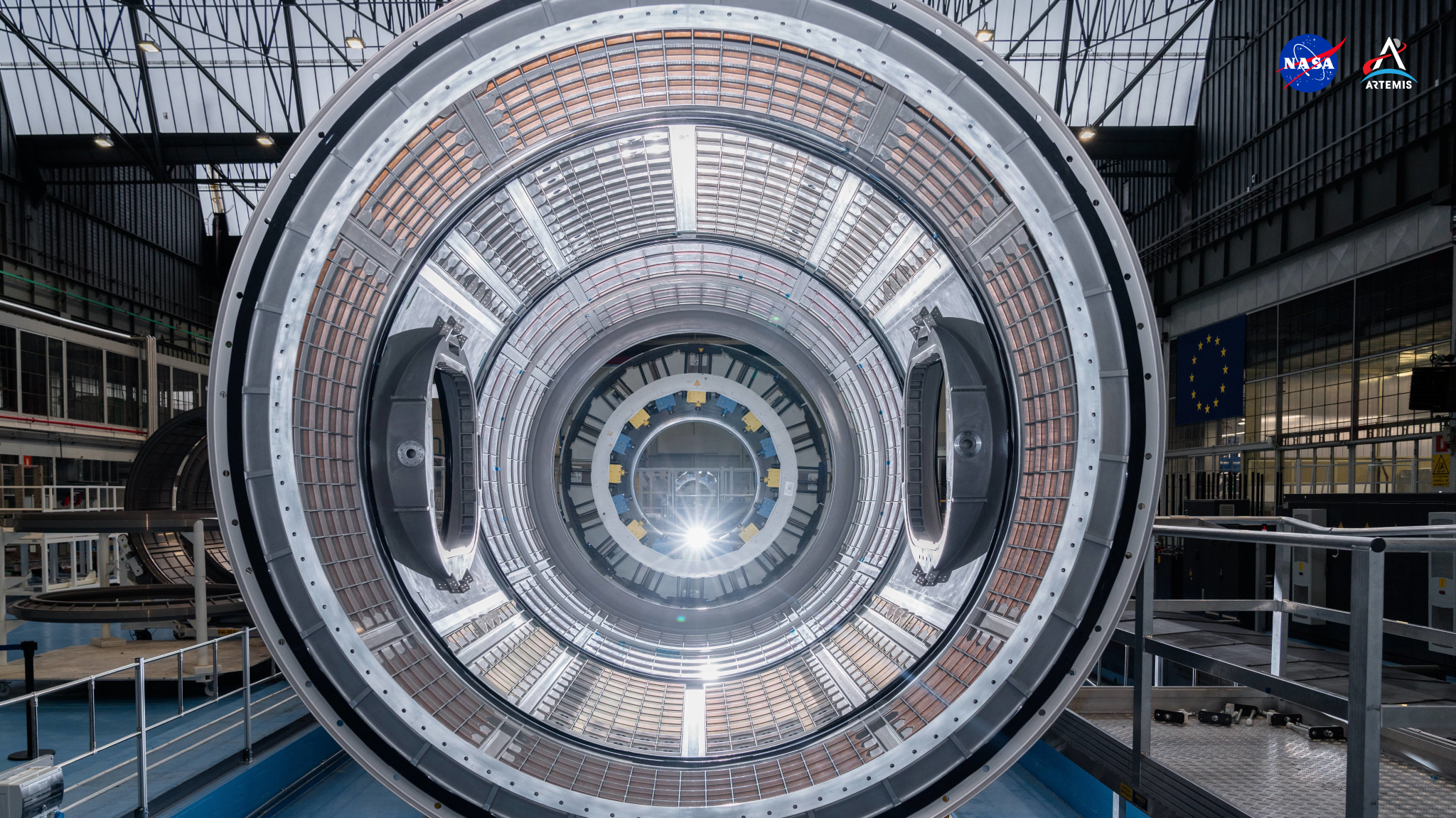 Dominating the frame of this background is the Gateway space station’s HALO (Habitation and Logistics Outpost) module, a giant silver-colored metal tunnel formed of segments welded together. A light source at the end of the tunnel shines toward the viewer, illuminating a grid-like pattern within the metallic sections. At each side, two large portholes open to the outside. HALO is seen here within a large building in Turin, Italy, where it was welded and tested.