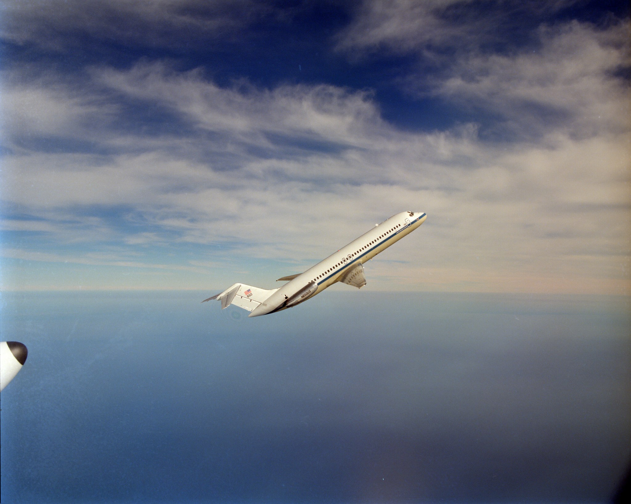 A white DC-9 plane with a blue stripe and text that says “Lewis Research Center” flies upward against a blue sky with several wispy white clouds.