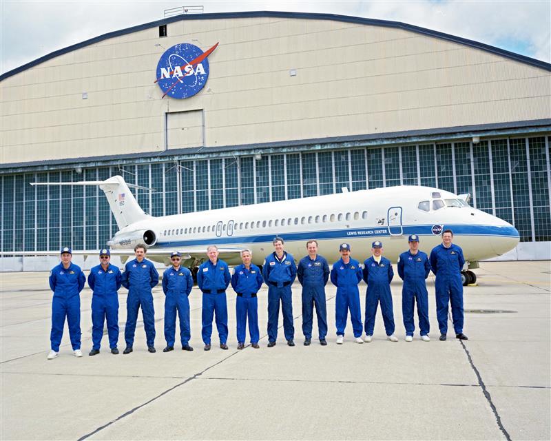 Twelve people wearing blue NASA flight suits stand in front of a white plane with a blue stripe and text that says, “Lewis Research Center.” In the background is the tan NASA Lewis hangar with a large NASA meatball logo sign affixed to the face of the building.