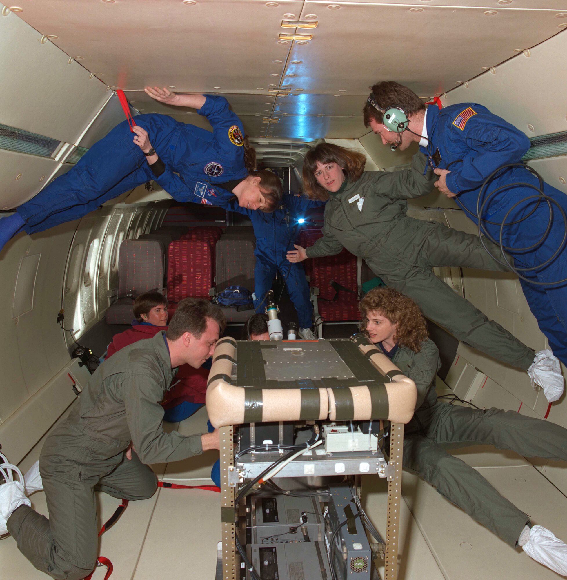 Three people wearing green flight suits and four people wearing blue flight suits float around a plane cabin and interact with experiment equipment.