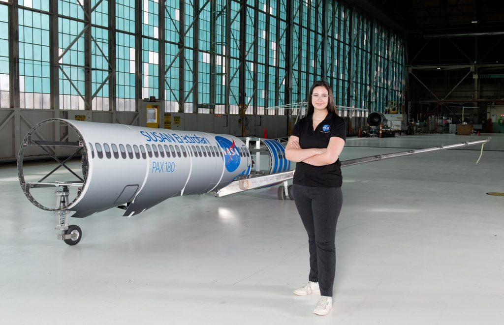 A woman wearing a black polo shirt with the NASA insignia, dark gray pants, and white shoes stands inside an aircraft hangar in front of a scaled aircraft model. The words “SUSAN Electrofan” and “PAX 180” are printed on a graphic along with fuselage, with a large NASA insignia in the center of the plane. The rear of the aircraft model, along with the wings and tail, are taken apart to display the inner components that make up its electrified propulsion system.