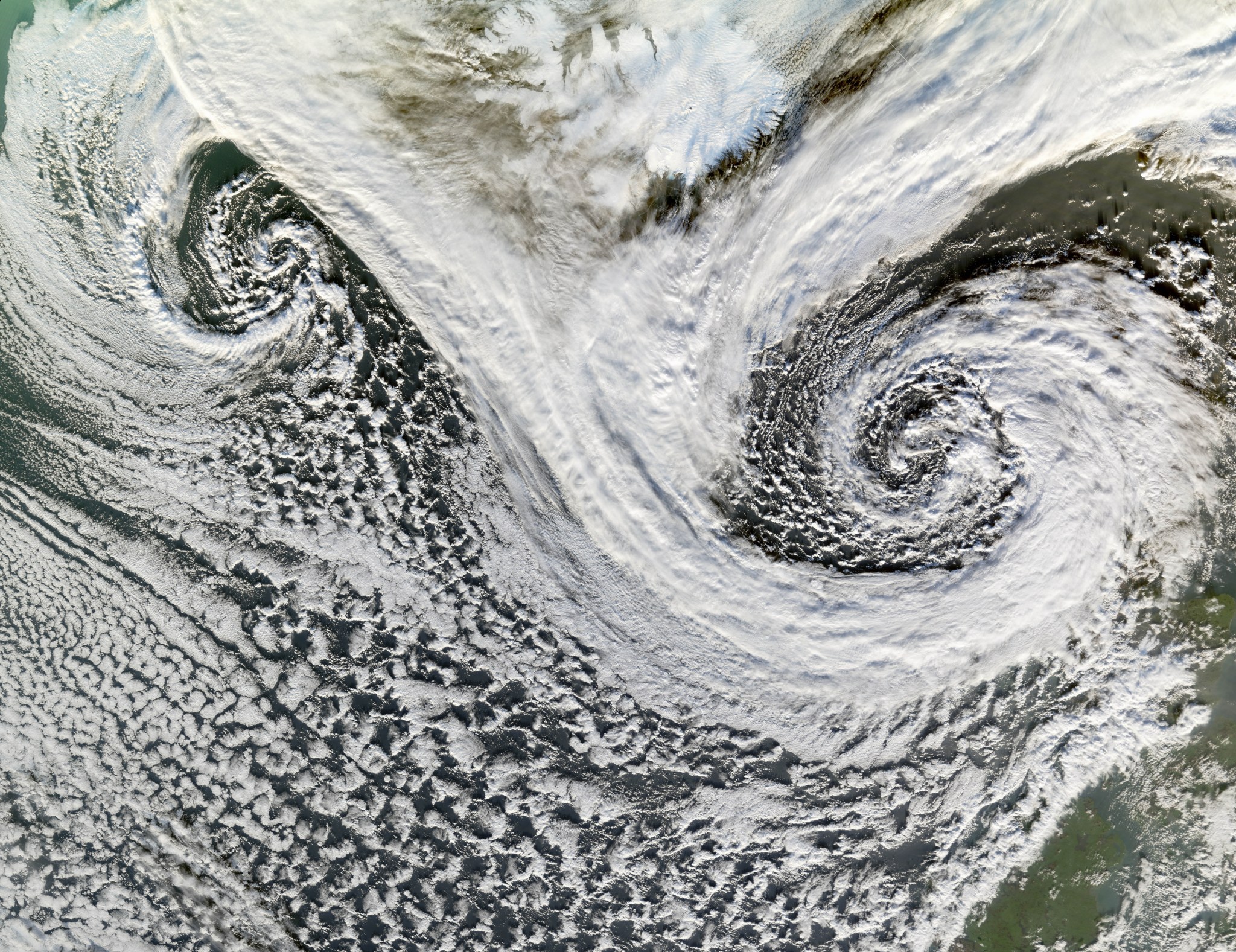 Two cyclones swirl south of Iceland in this satellite image. While the two storms' distinctive spiral patterns are clearly visible at left and right, white clouds cover most of the image. Some of Iceland can be seen at the top middle of the image, as well as part of Scotland at bottom right.